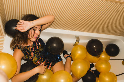 From above of young female in festive dress sleeping in bathtub filled with colorful balloons after holiday celebration