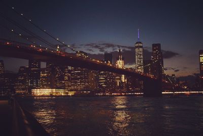 Illuminated brooklyn bridge over east river against city skyline against sky at dusk