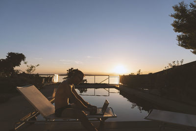 Side view of shirtless man using laptop while sitting by swimming pool against sky during sunset