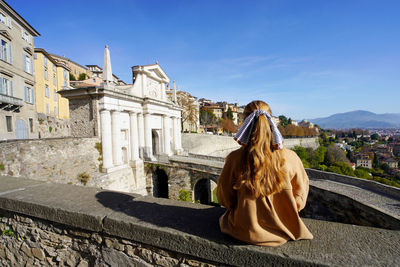 Young woman sitting on bergamo wall looking at porta san giacomo gate and venetian works of defence