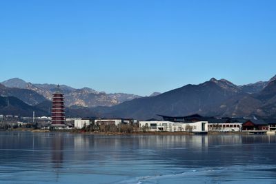 Tower at lakeshore by mountains against clear blue sky