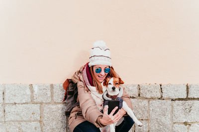 Cheerful woman taking selfie with dog against wall