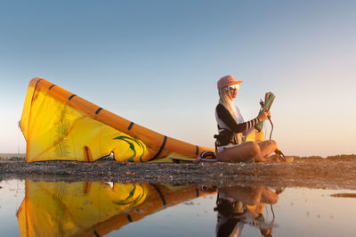 Attractive stylish young caucasian woman in cap sunglasses and kitesurfer outfit sits on the sandy