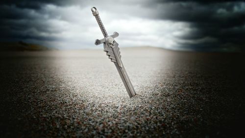 Sword in sand at beach against cloudy sky