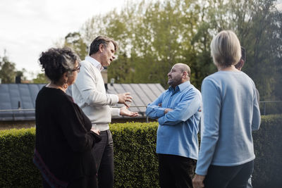 Mature man gesturing while talking to friends by hedge in yard