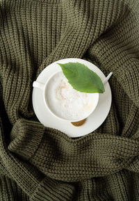 Cup of cappuccino coffee on green sweater top view flat lay