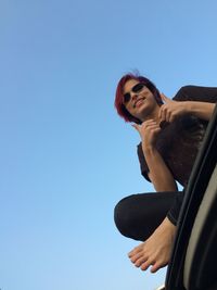 Low angle portrait smiling young woman showing thumbs up against clear sky