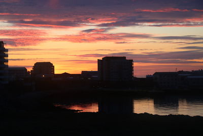 Silhouette buildings by river against orange sky