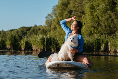 Happy young woman on the lake at early morning sitting on the sup board with her dog