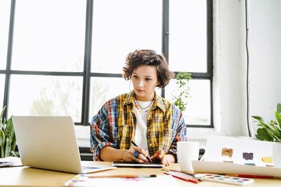 Young graphic designer using laptop sitting in front of window at desk