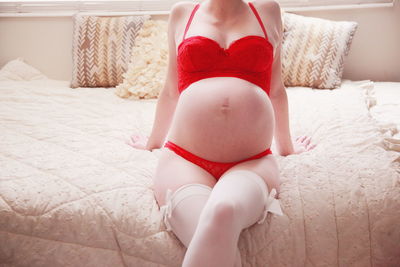 Midsection of pregnant woman in red bikini sitting on bed at home