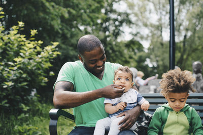 Smiling father feeding baby boy while sitting with son on bench at park