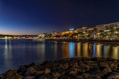 Illuminated buildings by sea against sky at night