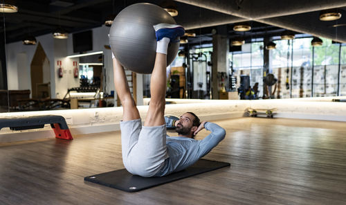 Bearded adult male athlete holding legs on fit ball and doing abdominal crunches on mat during fitness training in gym