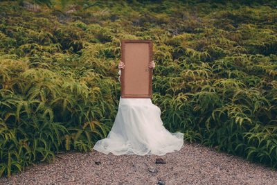 Bride holding empty picture frame while sitting by plants on field