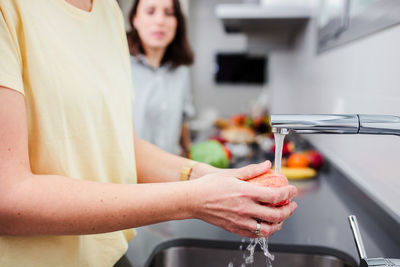 Midsection of daughter washing apple while standing by mother in kitchen at home