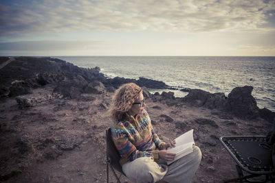 Woman reading book while sitting at beach against sky