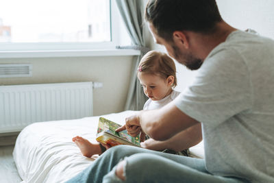 Happy father and baby girl little daughter having fun reading a book in children room at home