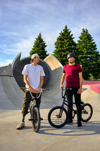 Smiling male riders with bmx bikes standing against sky at skateboard park