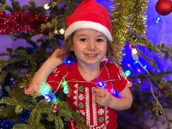 Portrait of cute girl wearing santa hat standing against christmas tree
