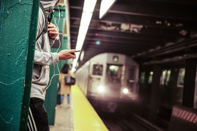 Midsection of man listening music while standing by train at railroad station platform