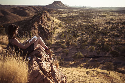 Side view of woman reading book while sitting on rock in desert