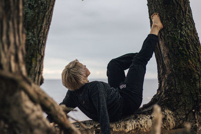 Rear view of woman sitting on tree trunk at beach against sky