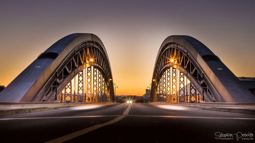 Illuminated road by bridge against sky during sunset