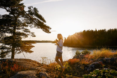 Full length of woman talking on mobile phone while standing at lake against sky during sunset