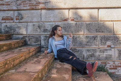 Young woman sits on grey stairs looks away dressed in ripped black jeans and grey upper. lifestyles.