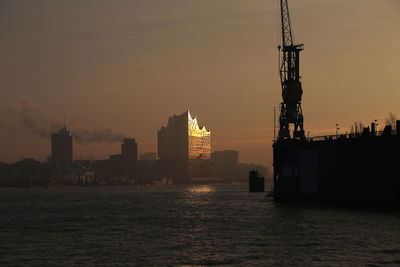 Silhouette buildings by sea against sky during sunset