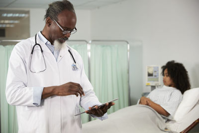 Male doctor using tablet pc with patient on bed in background at hospital