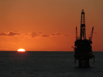 Silhouette oil industry in sea against sky during sunset