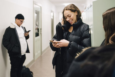 Smiling teenage student using smart phone in corridor