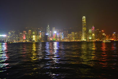 Illuminated buildings by river against sky at night