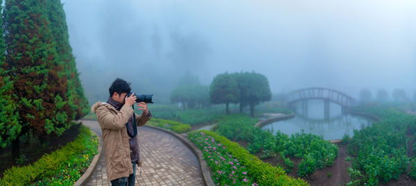 Side view of young man photographing with camera while standing in park during foggy weather