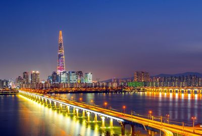 Illuminated modern buildings by river against sky at night