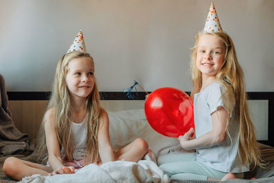 Happy children, girls, sisters, twins at home bed morning on their birthday festive caps red balloon