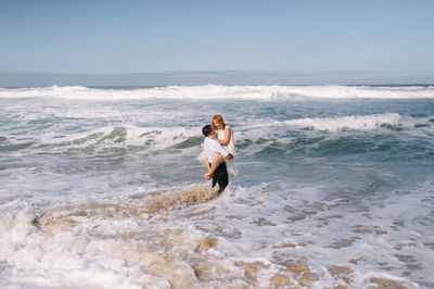 Man carrying woman while standing shore at beach against sky