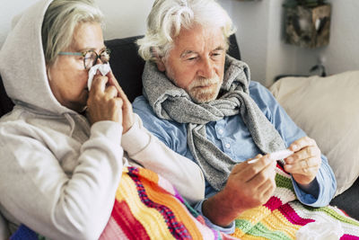 Close-up of senior man holding thermometer while sitting with wife on sofa at home