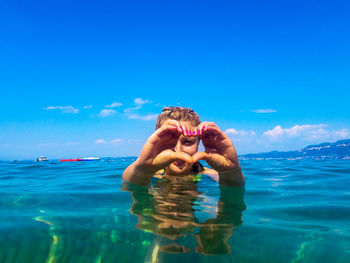 Woman making heart shape with hands in sea against blue sky during sunny day