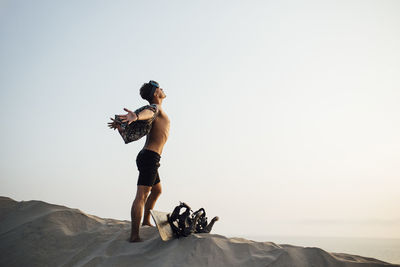 Young man with arms outstretched standing by sandboard in desert shorts at almeria, tabernas, spain