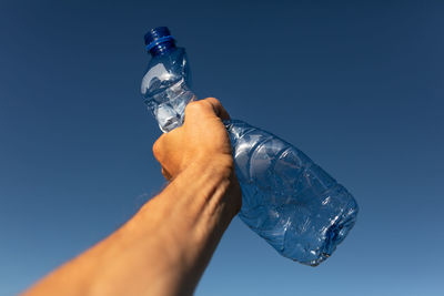 Cropped hand of person holding crumpled bottle against clear blue sky