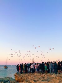 People by sea looking at balloons flying against sky during sunset