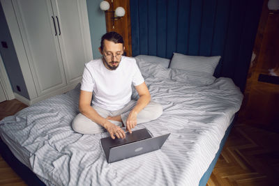 Man in a white t-shirt, a freelancer with glasses, sits on the bed at home with a laptop