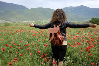Rear view of woman with arms outstretched walking amidst poppies on field against mountains