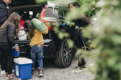 Family loading luggage in trunk while woman charging electric car