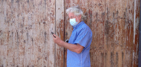 Side view of man wearing mask using mobile phone against wall