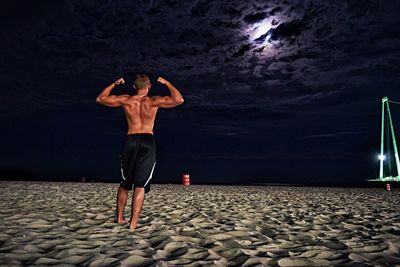 Rear view of shirtless muscular man standing at beach against cloudy sky during night