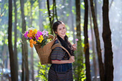 Portrait of beautiful young woman carrying flowers basket while standing in forest